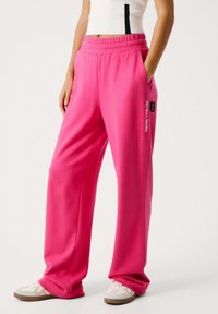 Lyserøde sweatpants med bred ben-design, elastisk talje, sidelommer og et "RED BULL RACING" logo på venstre side.