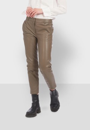 Persona vistiendo pantalones de cuero cortos color topo, botas negras gruesas y una camisa blanca de manga larga con las manos entrelazadas a la altura de la cintura.