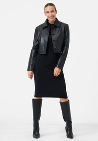 Zwarte leren cropped jas met vijf knopen, gecombineerd met een nauwsluitende zwarte jurk en zwarte laarzen tot de knie. Glad textuur en uitstraling.