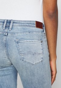 Jeans de denim azul claro con corte ajustado, que presentan un parche de cuero marrón en la cinturilla y un bolsillo trasero con costura en patrón de olas.
