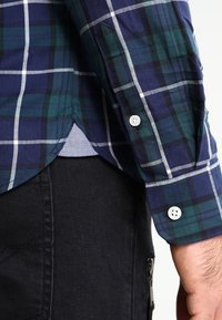 Camicia a scacchi con motivi blu, verdi e bianchi. Il materiale sembra morbido, con polsini abbottonati e un piccolo dettaglio sull'orlo.