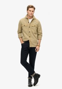 Chemise beige à boutons avec deux poches poitrine, associée à un jean en denim noir et des bottines noires. Montre portée au poignet gauche.
