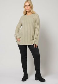 Beżowy sweter prążkowany z długimi rękawami, z wiązaniami przy mankietach, zestawiony z czarnymi dopasowanymi spodniami i czarnymi botkami za kostkę.