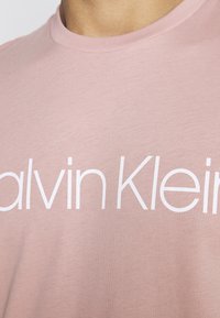 Jasnoróżowa koszulka z bawełny z okrągłym dekoltem i dużym białym napisem "Calvin Klein" na klatce piersiowej.