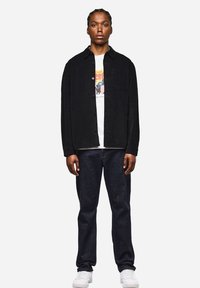 Giacca camicia in corduroy nero con bottoni frontali, taschino sul petto, indossata sopra una t-shirt bianca con grafica, abbinata a jeans scuri e sneakers bianche.