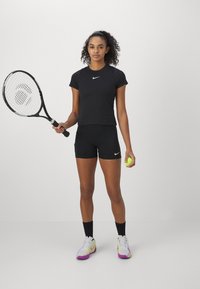 Top corto deportivo negro con un pequeño logo blanco de Nike, pantalones cortos negros y zapatillas blancas con acentos morados; sosteniendo una raqueta de tenis y una pelota.