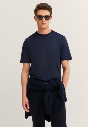 Man wearing dark sunglasses and a navy blue t-shirt with a navy sweater tied around his waist against a plain light background.