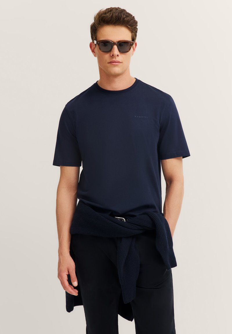 Mężczyzna w ciemnych okularach przeciwsłonecznych i granatowej koszulce z granatowym swetrem przewiązanym w talii na jasnym, jednolitym tle.