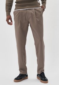 Khaki plisované kalhoty, lehký materiál, volný střih, kapsy po stranách, kombinované s černými teniskami a texturovaným hnědým svetr.