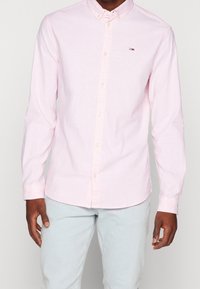 Mann in hellrosa Hemd mit Knöpfen und kleinem Logo auf der Brust, trägt hellblaue Jeans und steht vor einem hellgrauen Hintergrund.