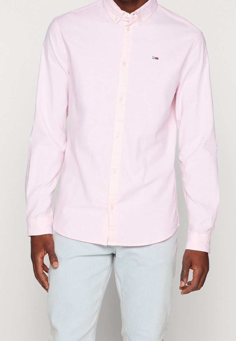 Mann in hellrosa Hemd mit Knöpfen und kleinem Logo auf der Brust, trägt hellblaue Jeans und steht vor einem hellgrauen Hintergrund.