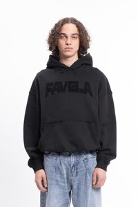 Jeune personne avec des cheveux bouclés portant un sweat à capuche noir avec le texte "FAVELA" et un jean bleu clair ample sur fond blanc.