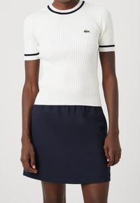 Bílé žebrované tričko s námořnicky modrým lemem u límce a rukávů, kombinované s jednobarevnou námořnicky modrou sukní. Na hrudi je malé zelené logo.