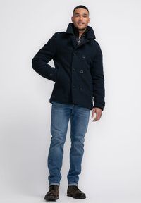 Abrigo peacoat de mezcla de lana azul marino con diseño de doble botonadura, grandes botones negros y bolsillos laterales. Llevado sobre una chaqueta acolchada.