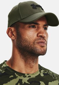 Gorra verde oliva con visera curvada, que presenta un logo negro en el frente. El material parece suave y tiene un diseño ajustado.