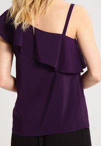 Fialový top na jedno rameno s výraznou volánkovou ozdobou kolem výstřihu, tenkými ramínky a hladkým, lehkým materiálem.