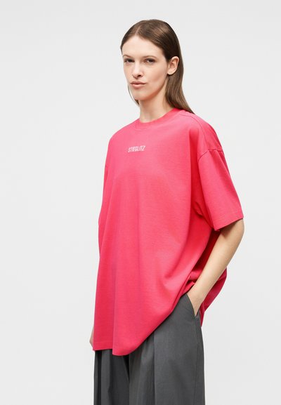 Γυναίκα με μακριά καστανά μαλλιά, φορούν χαλαρό φωτεινό ροζ T-shirt με το κείμενο "STIEGLITZ" και σκούρο γκρι παντελόνι με φαρδιά μπατζάκια, στεκόμενη με το ένα χέρι στην τσέπη.