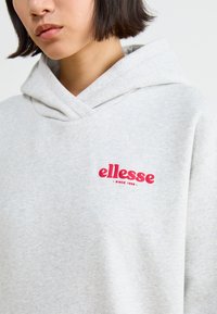 Szürke kapucnis pulóver elülső zsebekkel és húzózsinórral. A mellkasán piros "ellesse" logó található. Puha, texturált anyag.