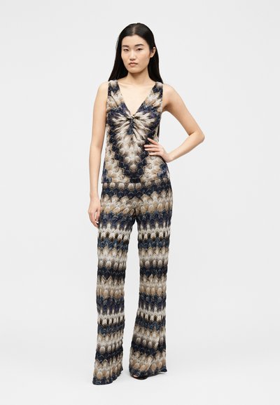 Γυναίκα με μακριά σκούρα μαλλιά που φοράει ένα αμάνικο ολόσωμο με V-λαιμόκοψη και πολύχρωμο ζικ ζακ μοτίβο σε αποχρώσεις του μπλε, μπεζ και μαύρου.