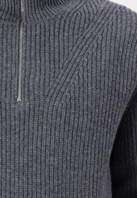 Grauer Wollpullover mit Reißverschluss bis zum Hals, vertikaler Rippstruktur, silbernem Reißverschluss und hohem Kragen. Zartes geometrisches Muster auf der Brust.