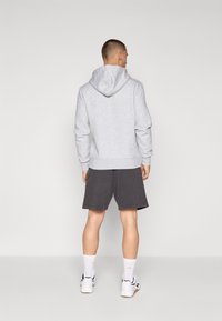 Γκρι φούτερ με κουκούλα και χαλαρή εφαρμογή, συνδυασμένο με σκούρες βερμούδες. Μοντέλο που φοράει λευκά αθλητικά παπούτσια και κάλτσες μέχρι τον αστ�ράγαλο, γυρισμένο μακριά από την κάμερα.