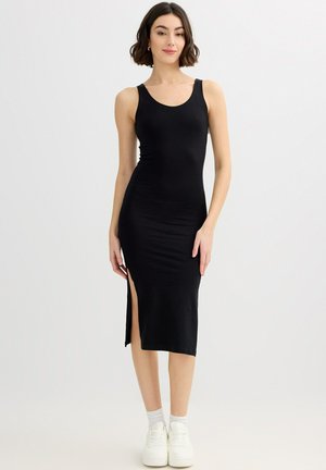 Mujer con un vestido midi negro sin mangas ajustado con una abertura lateral, calcetines blancos tobilleros y zapatillas blancas, de pie frente a un fondo liso.