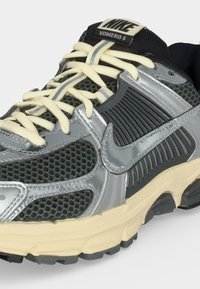 Αθλητικό παπούτσι Nike Vomero 5 σε γκρι και μαύρο με μπεζ κορδόνια και αντανακλαστικό ασημένιο λογότυπο Nike στην πλευρά.