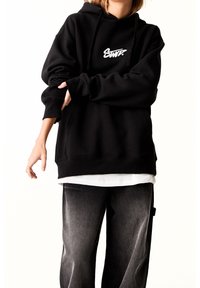 Personne portant un sweat à capuche noir avec un logo blanc, un t-shirt blanc en-dessous et un jean noir délavé, les bras croisés devant un fond blanc.