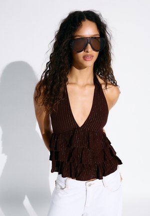 Žena s dlhými kučeravými vlasmi, ktorá nosí veľké tmavé slnečné okuliare, čierny vrchný diel s hlbokým V-výstrihom a volánmi bez rukávov a biele nohavice na jednoduchom pozadí.