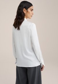 Biela, dlhá košeľa s dlhým rukávom vyrobená z ľahkej, hladkej tkaniny. Obsahuje golier a gombíkové manžety. Mäkko splývavá s voľným strihom.