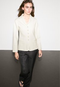 Junge Frau in einem cremefarbenen Knopfblazer, schwarzen Hosen und schwarzen Flip-Flops, die vor einem zweifarbigen weißen und grauen Hintergrund steht.