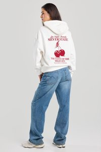 White hoodie with red graphic, featuring cherries and text on back. Paired with light blue loose-fit denim jeans and beige sneakers.
