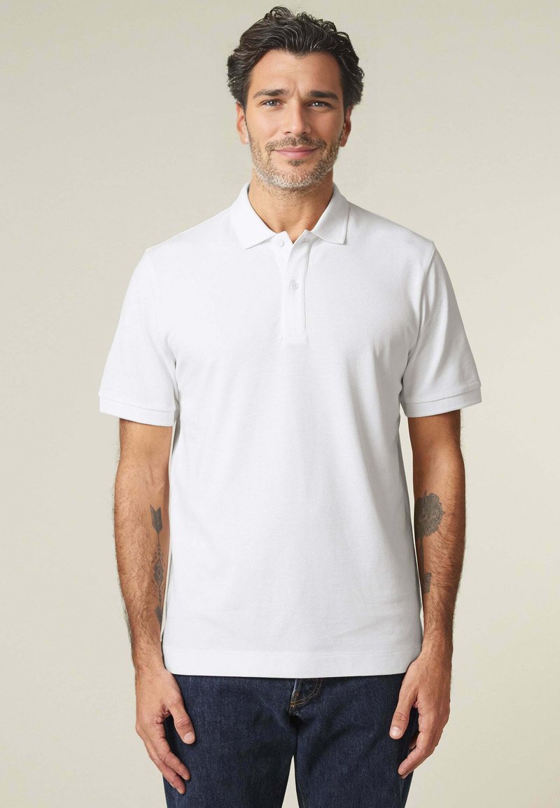 Witte polo met korte mouwen, klassieke kraag en een placket met drie knopen. Geko Abd met donkere denim jeans, met zichtbare arm tatoeages.