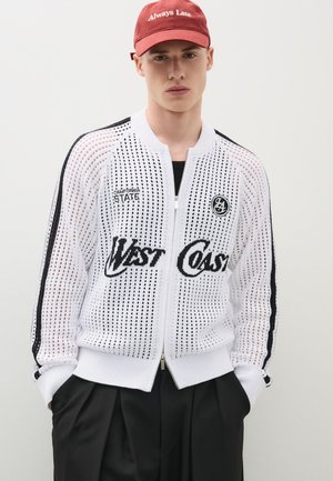Junger Mann mit roter Kappe und weißer Netz-Zipjacke mit schwarzem Text "West Coast" und "California State" über schwarzen Hosen.