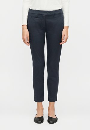 Γυναίκα που φοράει στενά μπλε παντελόνια, λευκό μακρυμάνικο τοπ και μαύρα φλατ παπούτσια, στέκεται μπροστά σε απλό ανοιχτό φόντο.