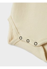 Beige baby bodysuit hoek met drie zwarte drukknoopjes en geschulpte stiksels op een witte achtergrond.