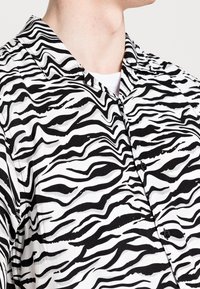 Camicia a maniche corte bianca e nera con un motivo a strisce zebrate ondulate. Presenta un colletto a bottoni e bottoni neri. Tessuto leggero.