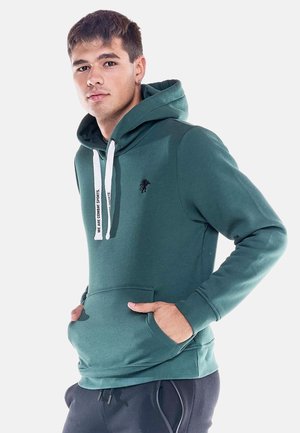 Jeune homme portant un sweat à capuche vert avec des cordons blancs et un pantalon noir, debout les mains dans la poche avant sur fond blanc.