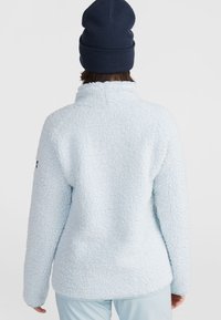 Lichtblauwe fleecetop met een gestructureerde, fuzzy buitenkant, hoge kraag en lange mouwen. Een marineblauwe beanie wordt op het hoofd gedragen.