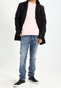 Mann in schwarzer Jacke über einem hellrosa Pullover, blauen Jeans und schwarzen Sneakern mit weißen Sohlen, der mit teilweise in den Taschen versenkten Händen dasteht.