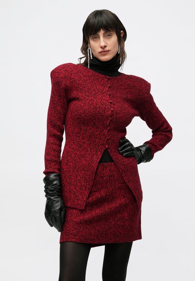 Červený textúrovaný pletený sveter s výraznými ramennými vypchávkami a gombíkovými detailmi, spojený so zodpovedajúcou sukňou, doplnený čiernymi rukavicami.