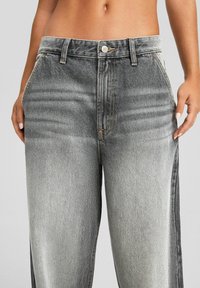 Nainen, jolla on yllä än korkeavyötäröiset vaaleanharmaat kulutetut farkut, joissa on etutaskut, vyölenkit ja nappikiinnitys, kädet lepäävät sivuilla.