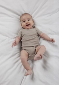 Bebeluș zâmbitor în combinezon bej, întins pe pat alb, cu brațele și picioarele ușor ridicate și relaxate.