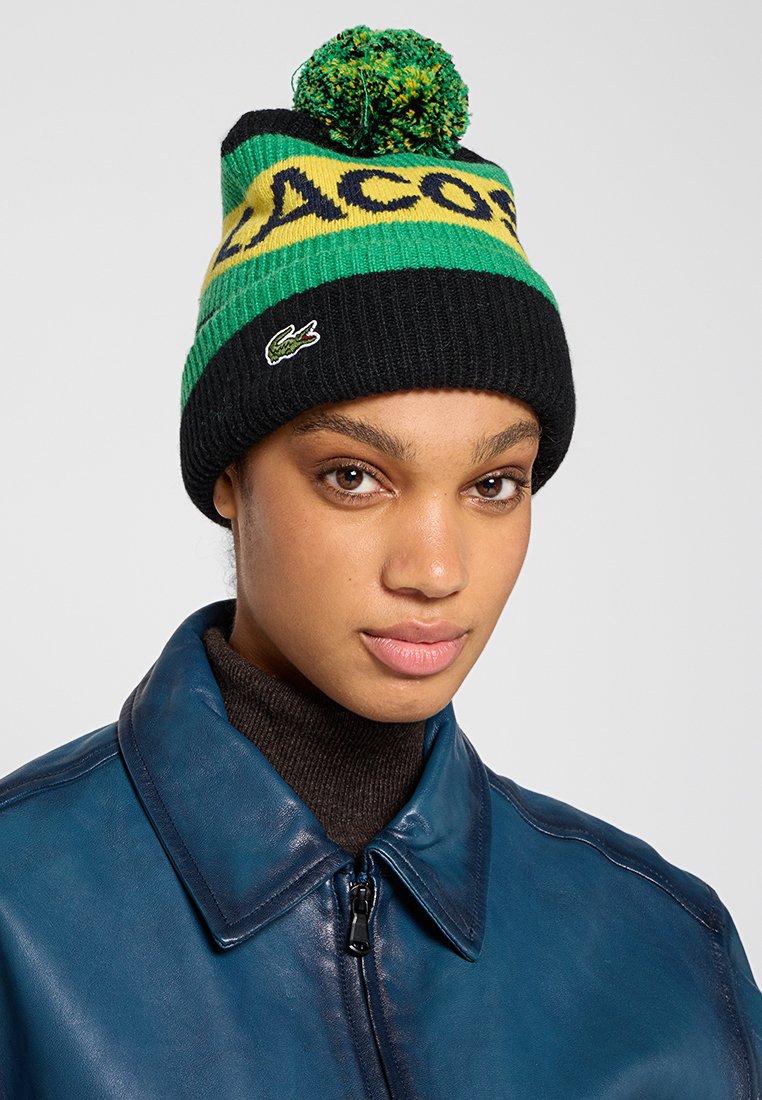 Zwarte gebreide beanie met groene en gele horizontale strepen, grote pompon bovenop en een klein groen krokodil logo.