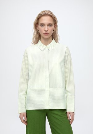 Femme aux cheveux ondulés clairs portant une chemise blanche boutonnée à fines rayures verticales et un pantalon vert, debout devant un fond uni.