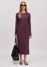 Robe à manches longues, de couleur violet profond, avec un détail en nœud à la taille. Le mannequin tient un sac fourre-tout noir en texture et porte des tongs noires.
