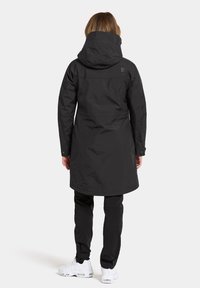 Schwarze, lange Jacke mit Kapuze, schlichtes Design und glatte Textur; getragen über schwarzen Hosen und weißen Schuhen.