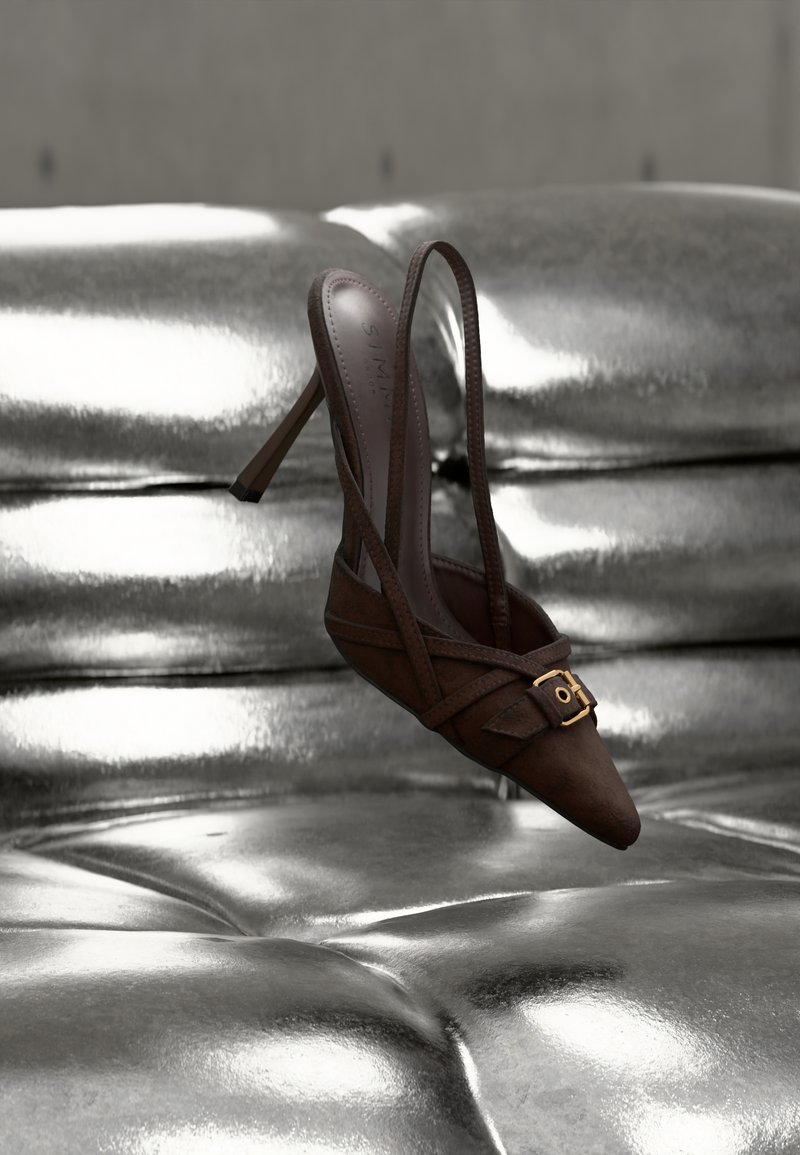 Καφέ καστόρινη γόβα με λουράκι πίσω, ψηλό τακούνι, μυτερή μύτη, σταυρωτά λουράκια και χρυσή διακόσμηση. Τοποθετημένη πάνω σε γυαλιστερή ασημένια επιφάνεια.