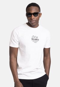 Weißes Baumwoll-T-Shirt mit kurzen Ärmeln, das mit schwarzem Text bedruckt ist, der "Aus DIESEM WELT" liest, sowie einem Logo. Getragen mit schwarzen Sonnenbrillen.