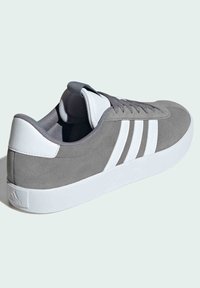 Zapatillas de ante gris con suela de goma blanca y tres franjas blancas. Lengüeta y tobillo acolchados, presenta una parte superior texturizada y suave.